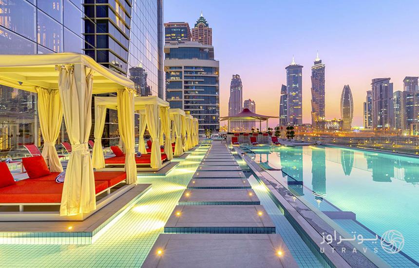 نمایی از استخری در یکی از هتل‌های دبی که درکنار آن چند تخت آفتابگیر قرمزرنگ با سایه‌بان کرمی قرار دارد.